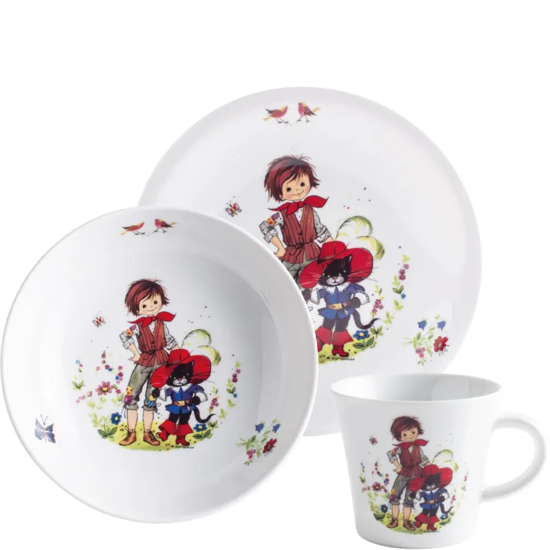 Drei Geschirrteile auf weißem Porzellan, darunter eine große flache Schüssel, ein flacher Teller und eine Tasse. Die Geschirrstücke sind mit bunten Illustrationen von einem Jungen in traditioneller Tracht und einem Hund dekoriert, umgeben von Blumen und Schmetterlingen.